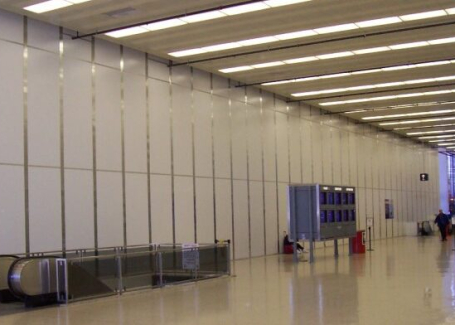 A large indoor space with high ceilings, featuring a long wall of white panels, overhead lighting, and an escalator leading down to another level.