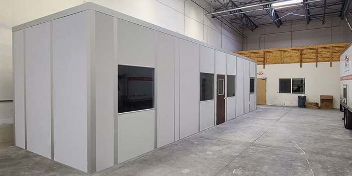 A freestanding modular office inside a warehouse, featuring a clean, white panel exterior with multiple windows and a single door. The structure is positioned on a concrete floor, with an open warehouse space surrounding it. Overhead lighting and exposed ceiling beams highlight the industrial setting.