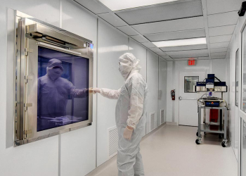 Technician in full cleanroom attire working inside an Allied Modular cleanroom with integrated pass-through chamber and wall-mounted equipment.