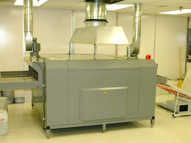 A specialized industrial machine inside a controlled workspace, featuring a large gray housing with integrated ventilation ducts extending upward. The machine includes a conveyor system on one side and appears designed for processing or manufacturing tasks. The enclosure ensures proper airflow and safety for the operation of the equipment.