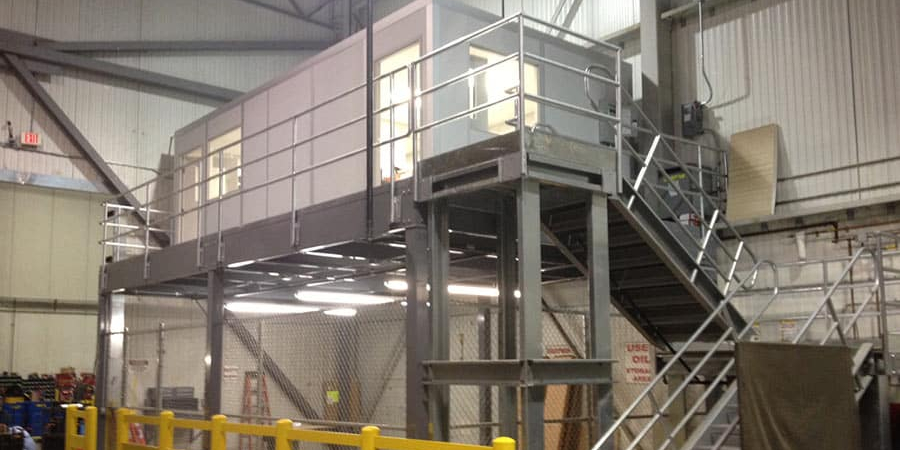 A modular mezzanine office space elevated above a workshop area with metal stairs and railings.