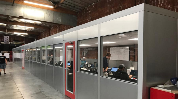 Long row of modular office spaces inside a warehouse, featuring individual workstations with glass windows and red-framed doors.
