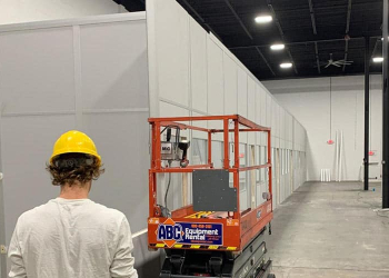 A man in a yellow hard hat stands next to orange scissor lift next to a modular office enclosure.