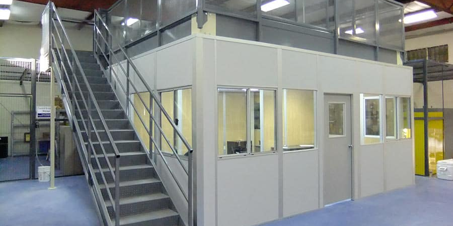 A mezzanine structure with a staircase leading to an upper level above an enclosed office space in a warehouse. The mezzanine is supported by metal beams and includes safety railings around the perimeter.