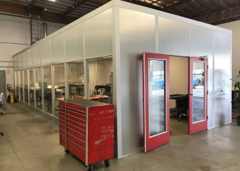 A modular office with glass windows and red doors installed inside a warehouse space.