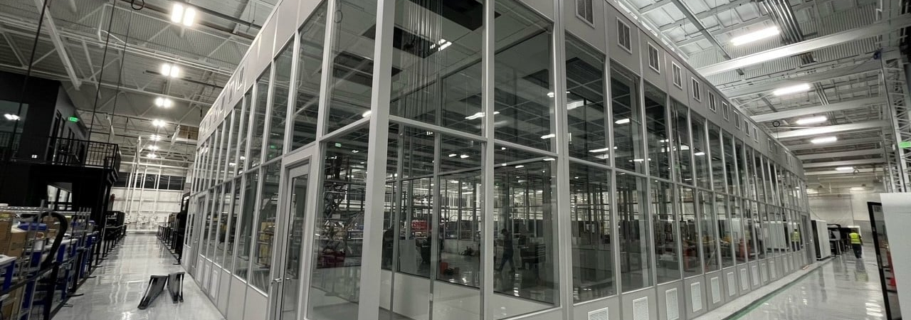 A modular factory structure with glass walls and a metal frame, located inside a large, well-lit industrial facility.