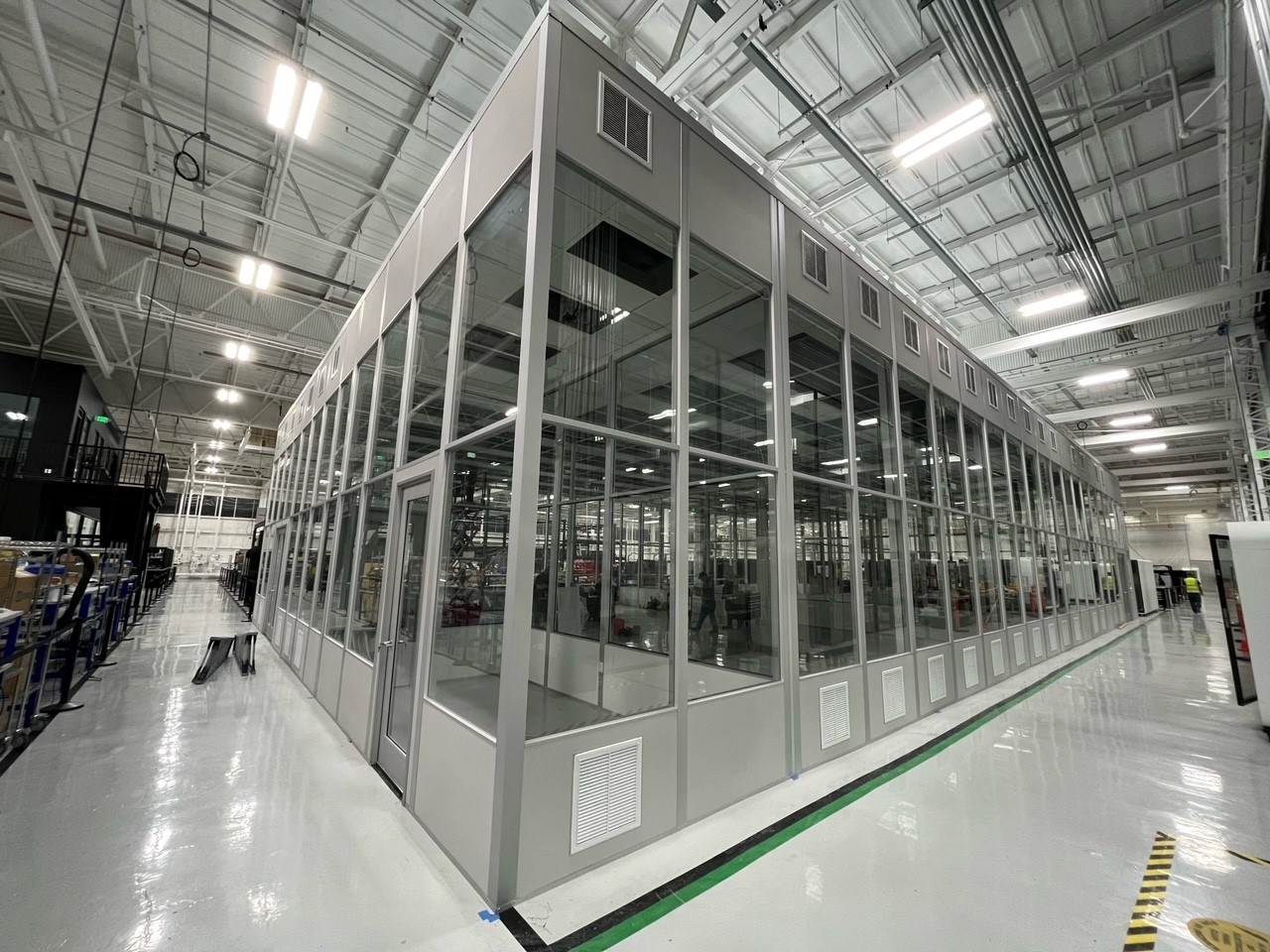 A modular factory structure with glass walls and a metal frame, located inside a large, well-lit industrial facility.