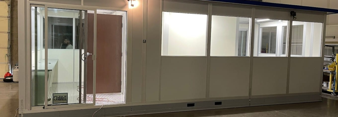 A modular office building with large windows and a door, assembled inside a warehouse.