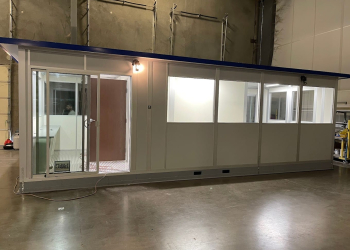A modular office building with large windows and a door, assembled inside a warehouse.