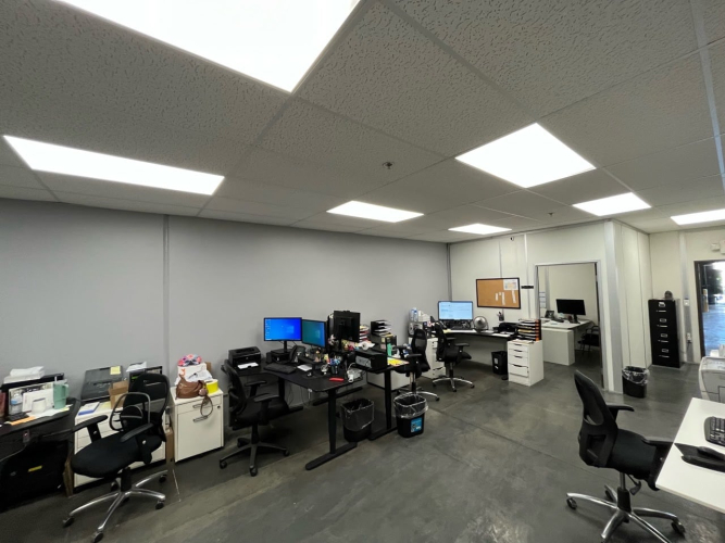 An office space with multiple desks and computer workstations, lit by overhead fluorescent lights.
