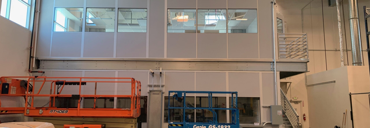 Two-story modular factory office with large windows and a staircase, situated inside an industrial warehouse, alongside scissor lifts for easy access and mobility.