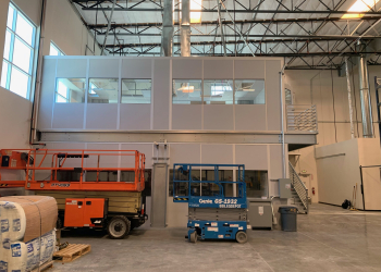 Two-story modular factory office with large windows and a staircase, situated inside an industrial warehouse, alongside scissor lifts for easy access and mobility.