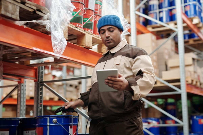 A warehouse worker using a tablet to manage inventory while standing next to neatly stacked shelves.