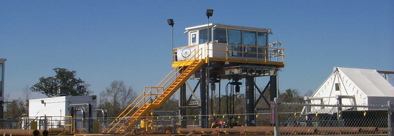 A tall modular observation tower in an industrial setting, featuring a white booth with large windows, supported by a steel structure with yellow stairs and railings.