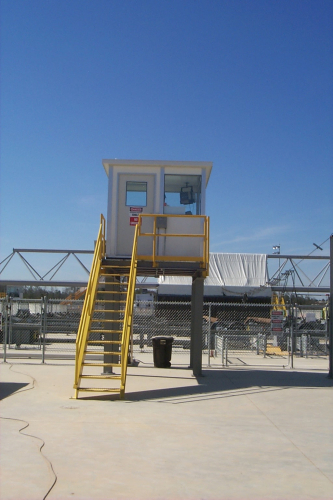 A modular observation tower with a white booth and yellow stairs, situated in an industrial area under a clear blue sky.