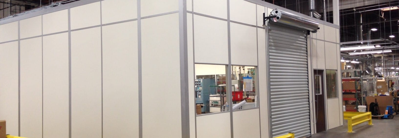 A prefabricated equipment enclosure inside a warehouse, featuring beige modular panels with metal framing. The enclosure includes a roll-up door, a window, and a standard entry door for access. Yellow safety barriers are positioned around the perimeter for protection. The enclosure is designed to house machinery or equipment securely within an industrial setting.