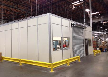 A prefabricated equipment enclosure inside a warehouse, featuring beige modular panels with metal framing. The enclosure includes a roll-up door, a window, and a standard entry door for access. Yellow safety barriers are positioned around the perimeter for protection. The enclosure is designed to house machinery or equipment securely within an industrial setting.