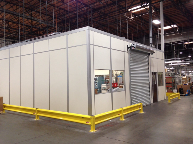A prefabricated equipment enclosure inside a warehouse, featuring beige modular panels with metal framing. The enclosure includes a roll-up door, a window, and a standard entry door for access. Yellow safety barriers are positioned around the perimeter for protection. The enclosure is designed to house machinery or equipment securely within an industrial setting.