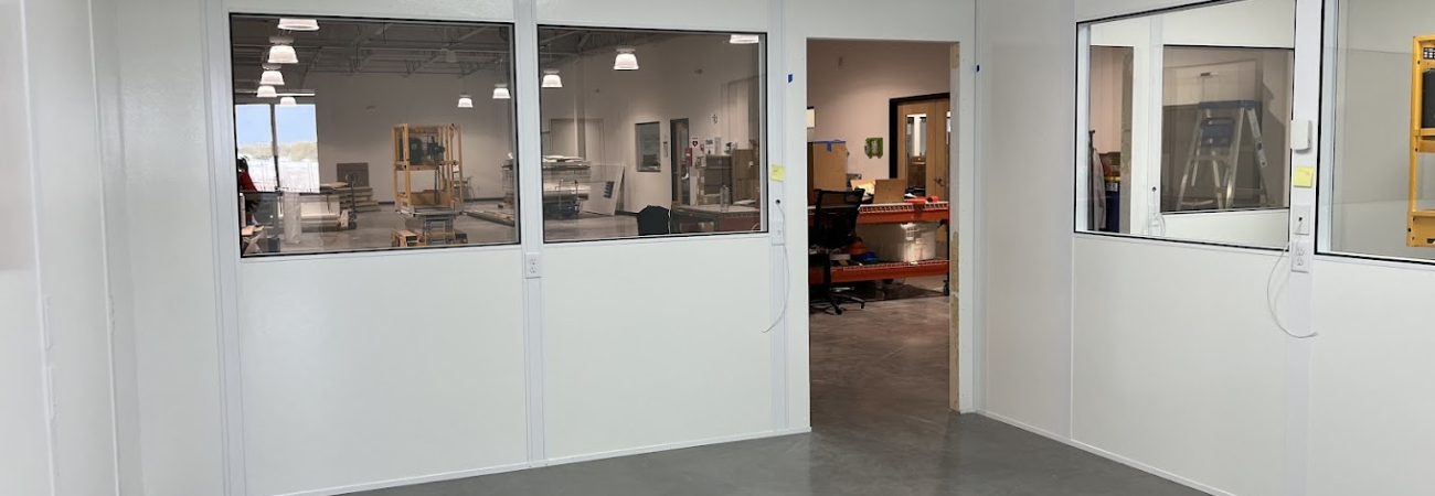 A clean, modern Allied Modular office inside a warehouse, featuring white panel walls, large windows for visibility, and an open doorway leading to the main warehouse area. The office has a suspended ceiling with recessed lighting and a polished concrete floor.