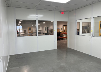 A clean, modern Allied Modular office inside a warehouse, featuring white panel walls, large windows for visibility, and an open doorway leading to the main warehouse area. The office has a suspended ceiling with recessed lighting and a polished concrete floor.