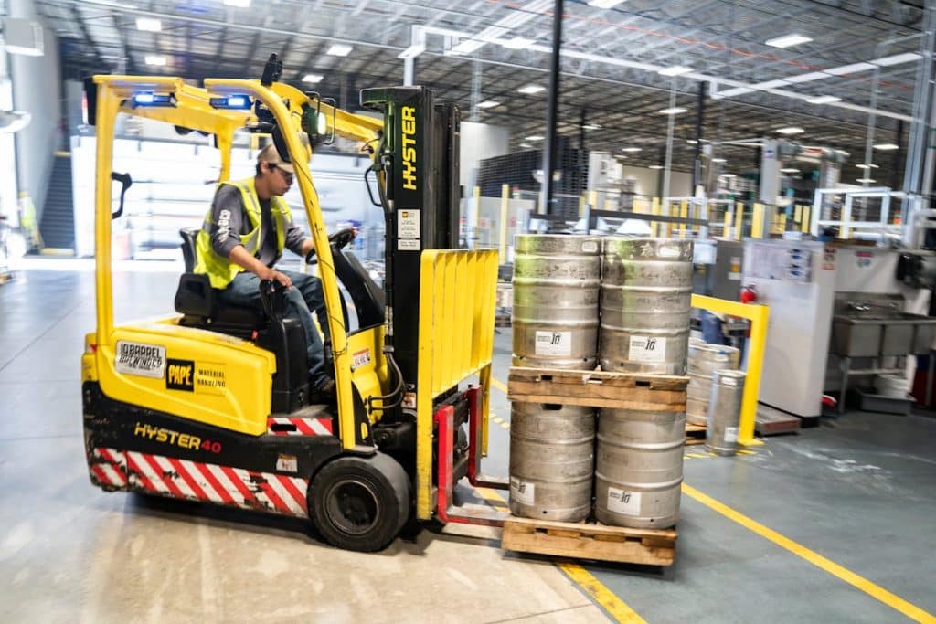 Warehouse worker operating a yellow forklift to move stacked metal barrels on a pallet.